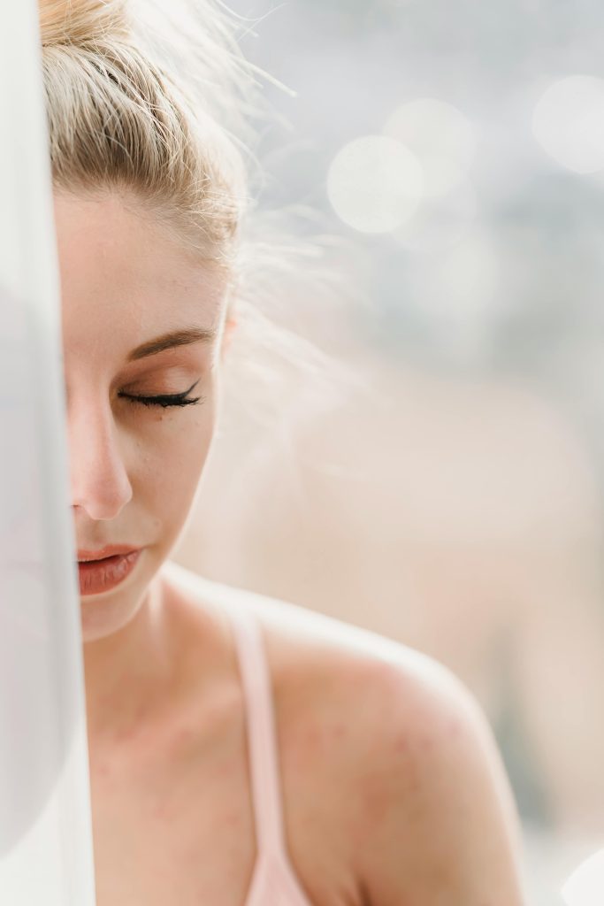 Young woman with eyes closed meditating near a window, enjoying peaceful sunlight.